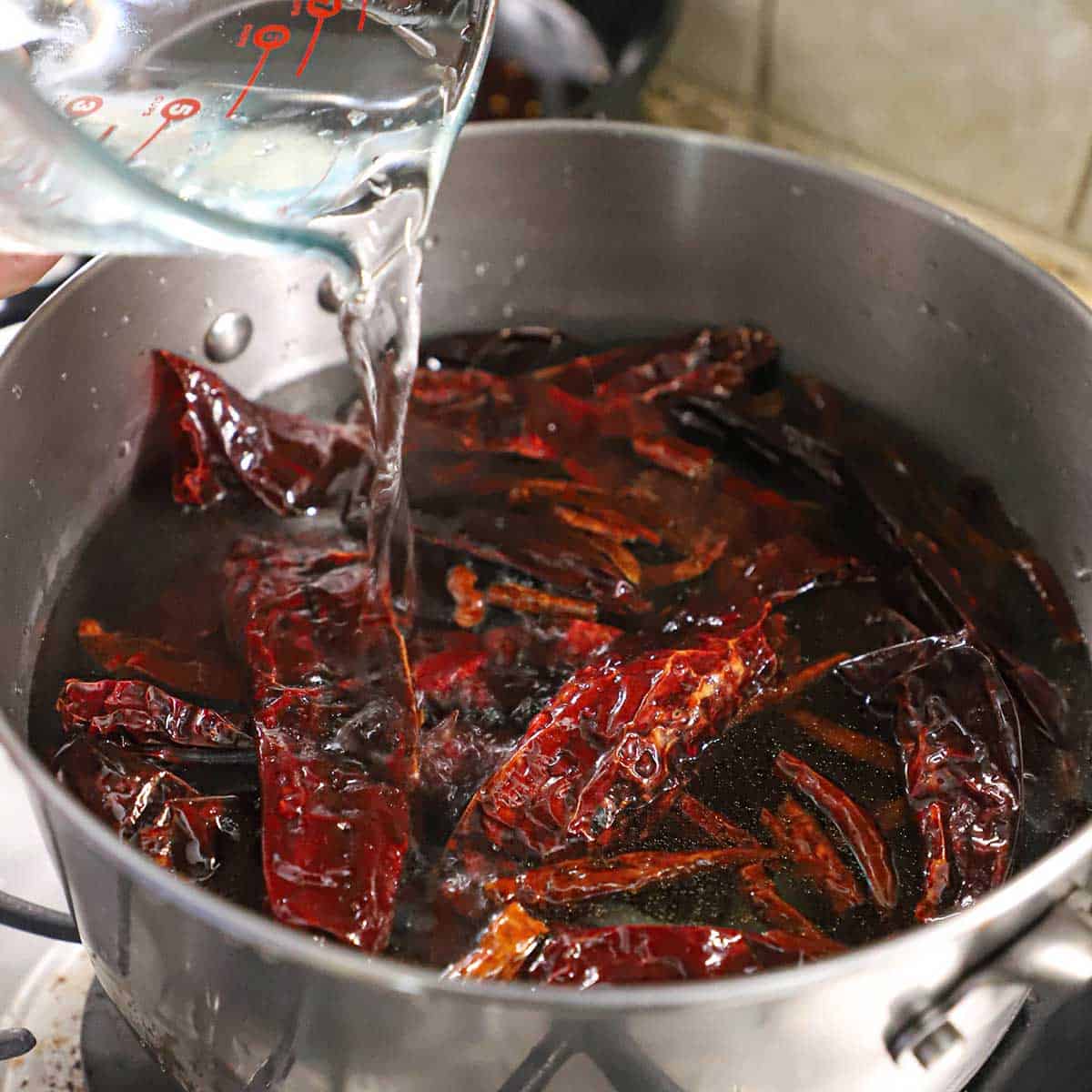 A person pouring water from a large glass measuring cup into a pot filled with dried chiles.