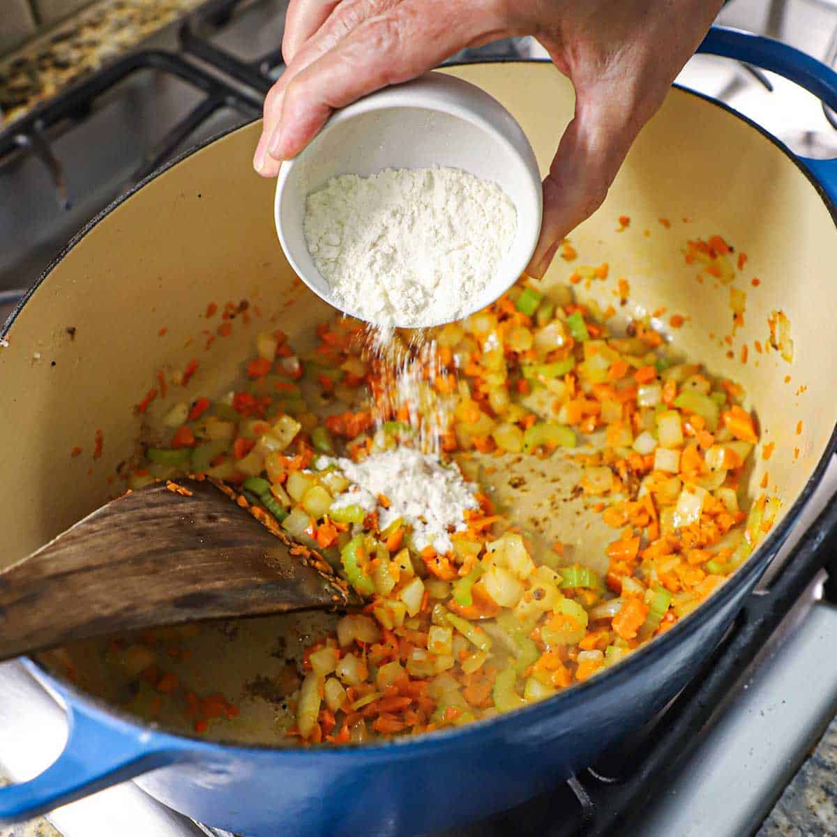 A person transferring a small mound of all-purpose flour into an oval Dutch oven that has chopped onions, carrots, and celery being sautéed in it.