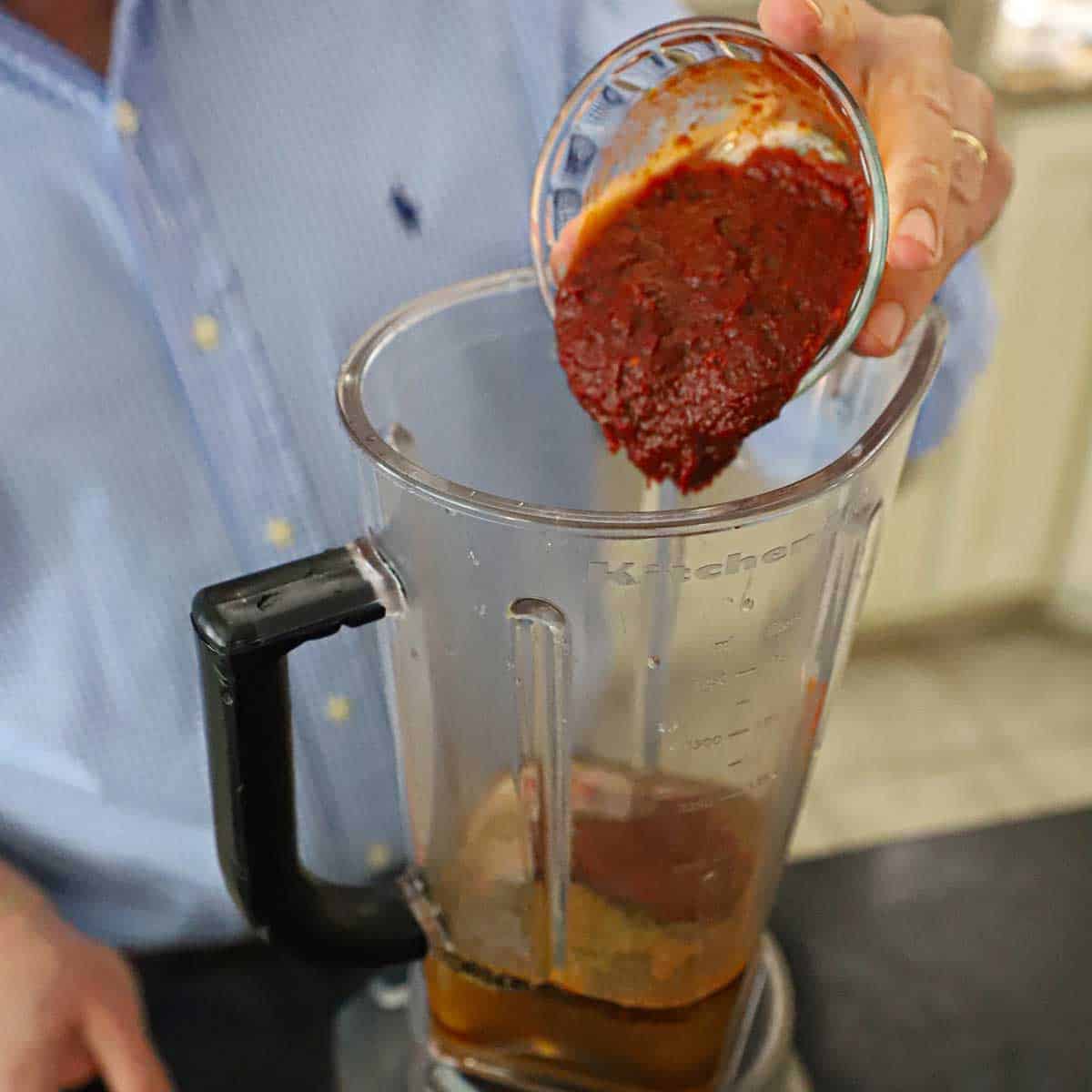 A person transferring homemade chili paste from a small glass bowl into a blender that contains broth and spices.