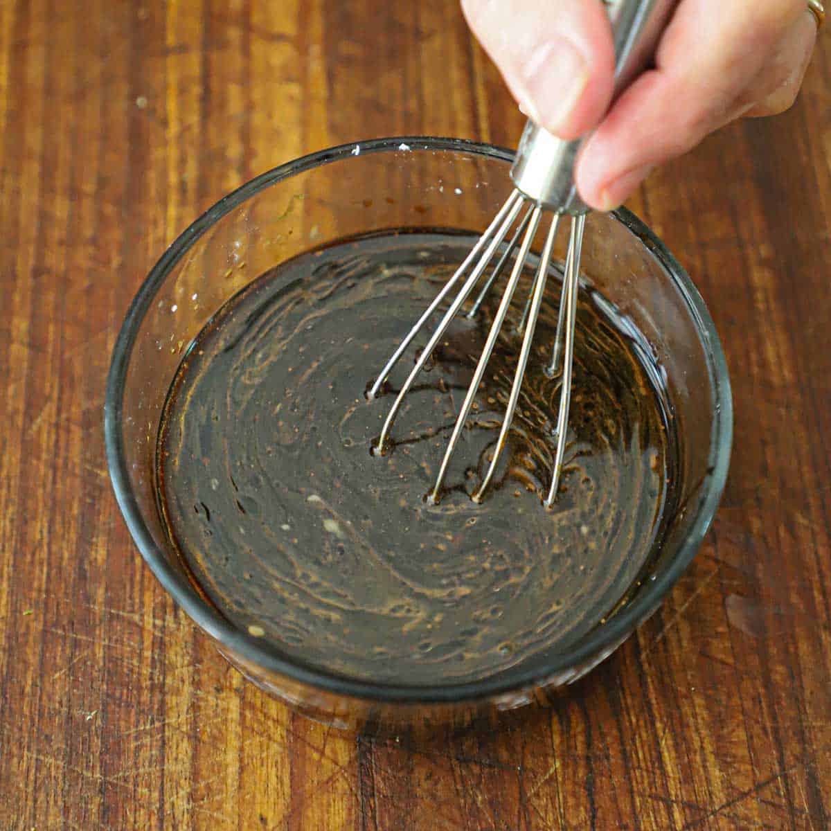 A person using a small whisk to mix a Chinese lo mein dark sauce in a small glass bowl on a wooden cutting board.