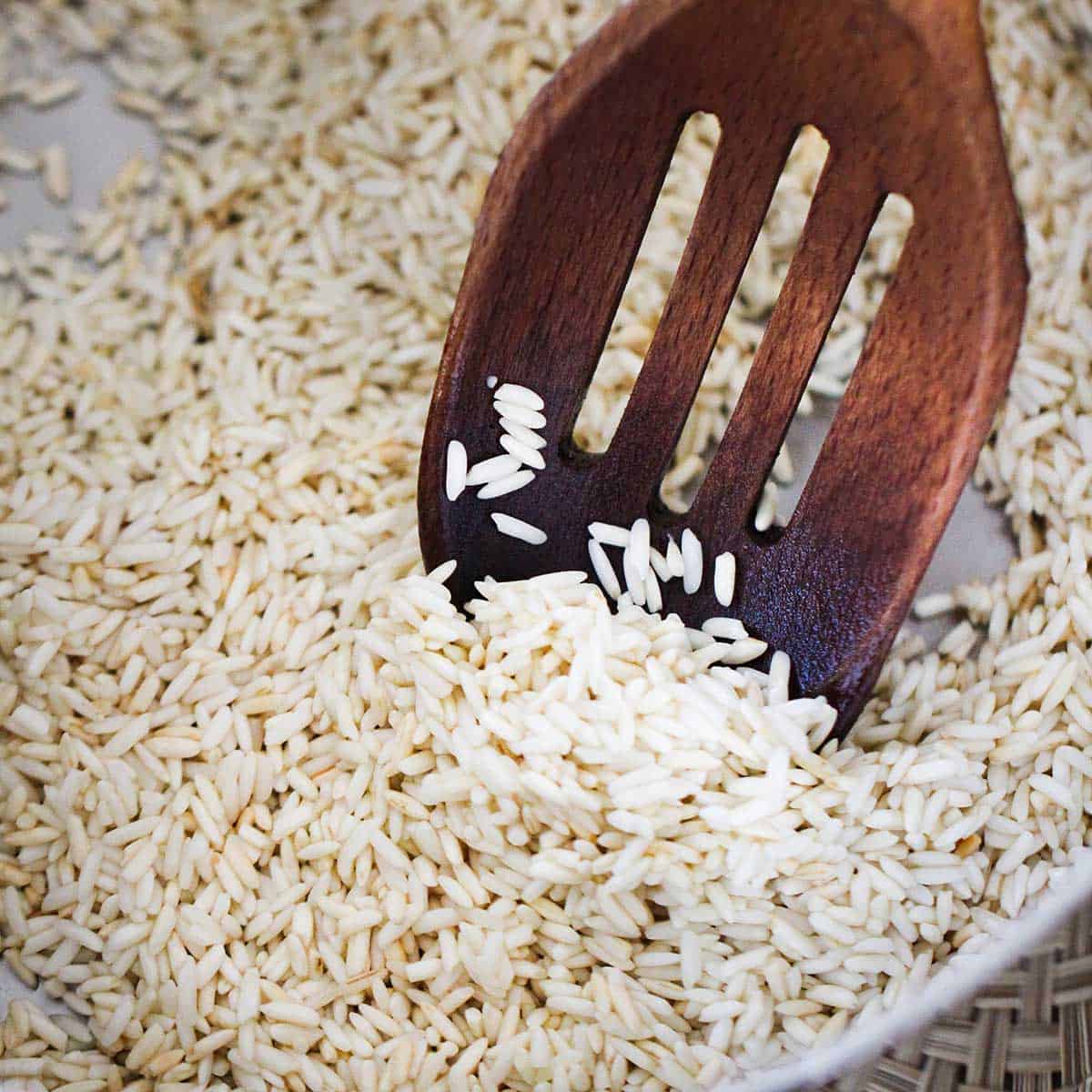 A wooden slotted spoon being used to stir long-grain rice in the bottom of a silver saucepan.