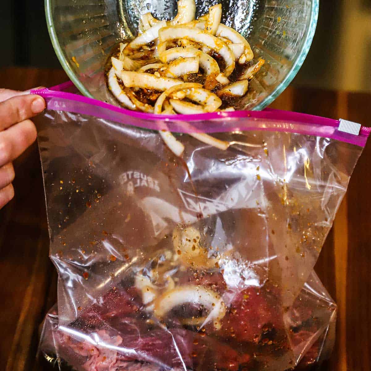 A person transferring a marinade and sliced onions into a plastic baggie that has an uncooked flat-iron steak in it.