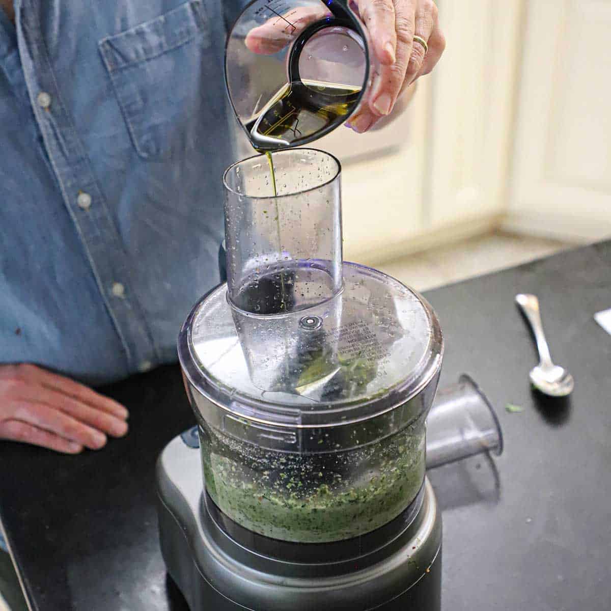 A person pouring extra-virgin olive oil through the tube of a food processor into the bowl that is processing pesto sauce.