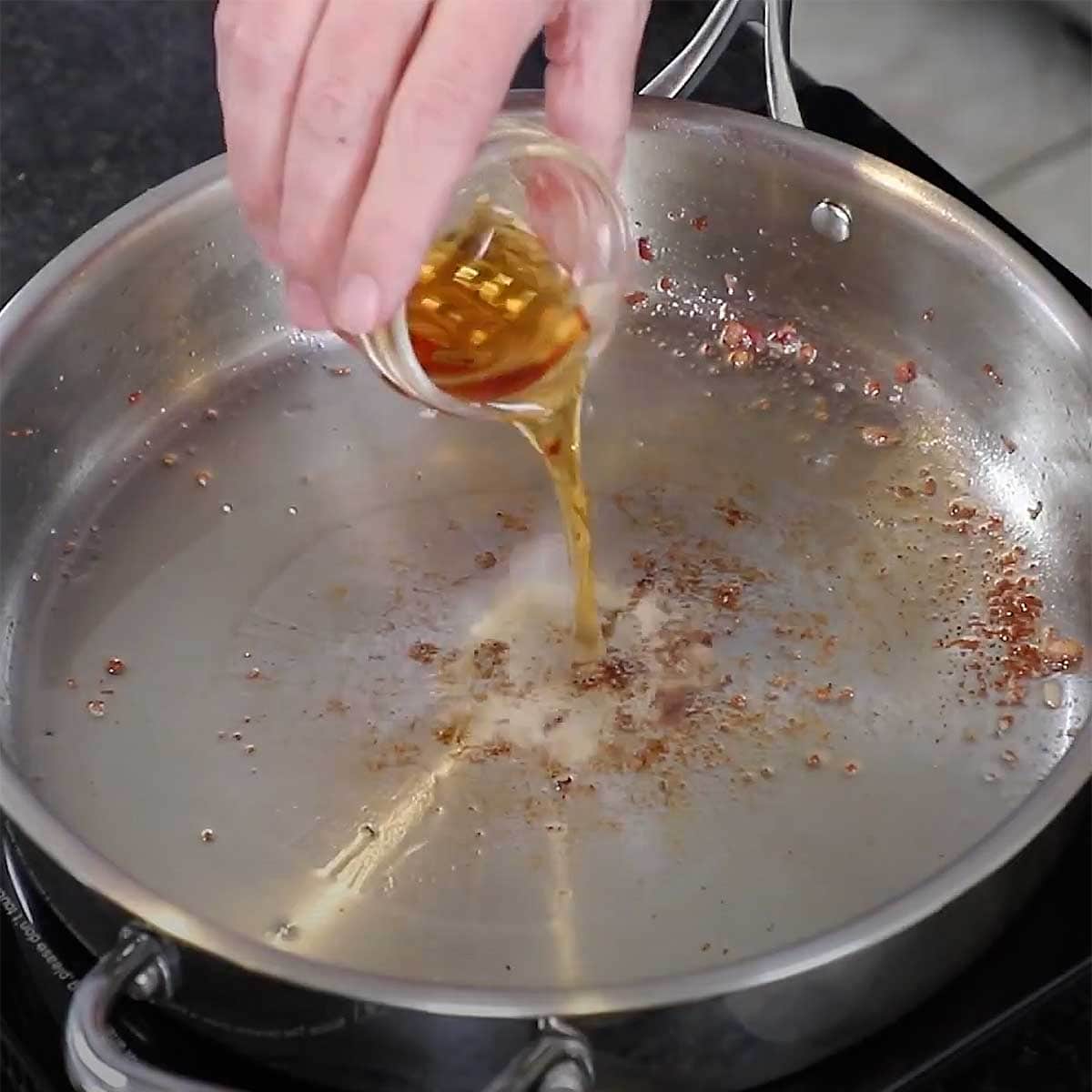 A person pouring Madeira wine from a small glass jar into a silver skillet on a stove top.