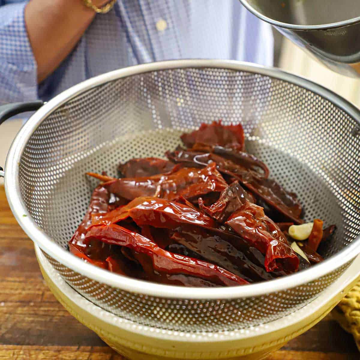 A colander resting on top of a ceramic bowl with a pile of dried chiles that have been rehydrated with boiling water.