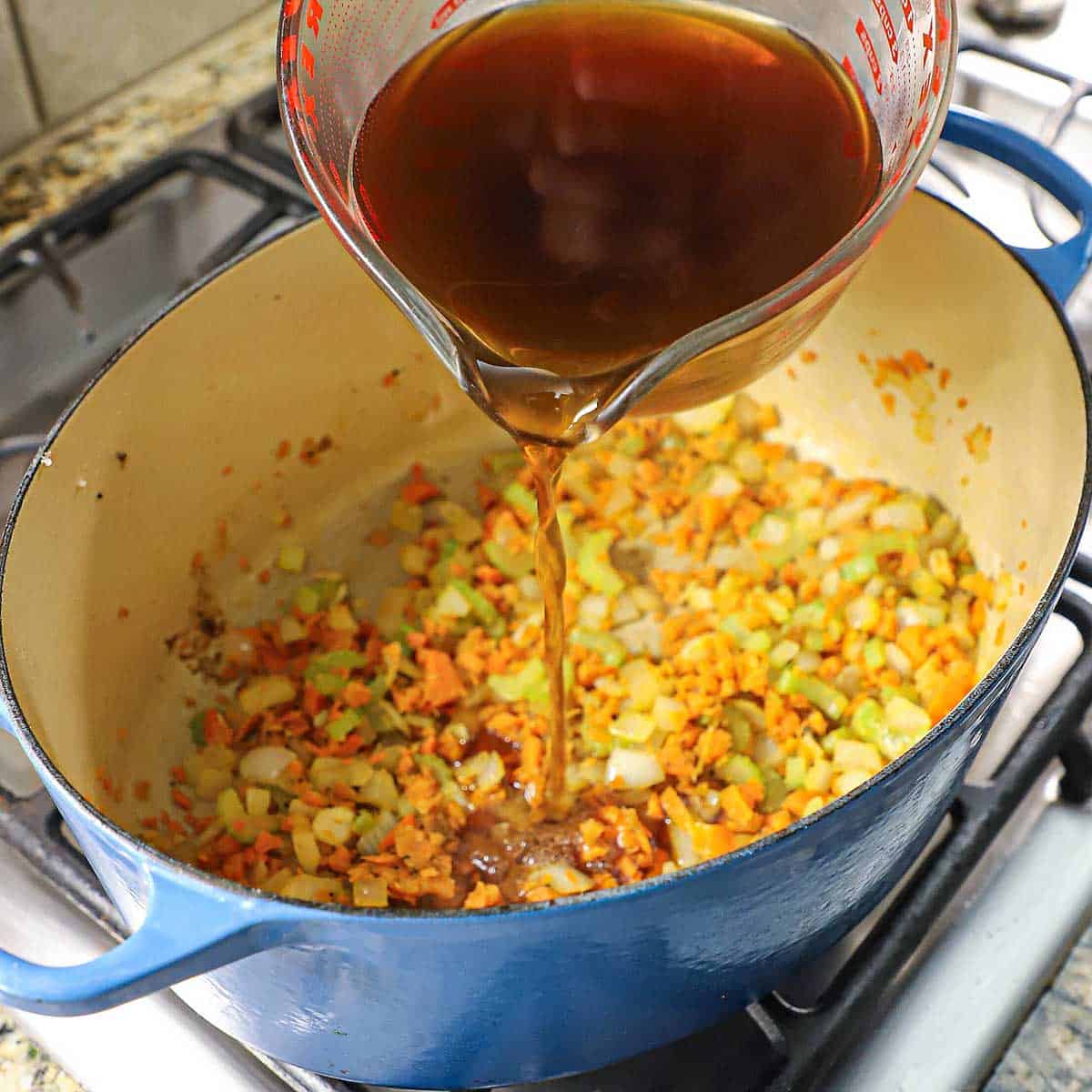 A person using a large glass measuring cup to transfer beef broth into a large oval Dutch oven filled with sautéed chopped onions, carrots, and celery.