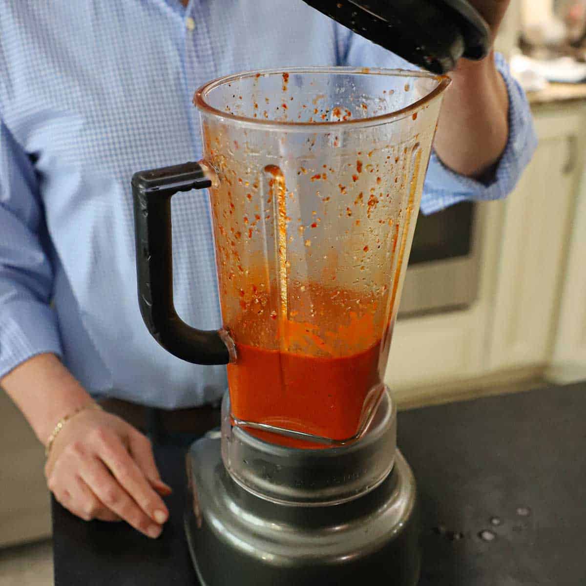 A person removing the lid from a blender that is half-filled with a puréed chili marinade.