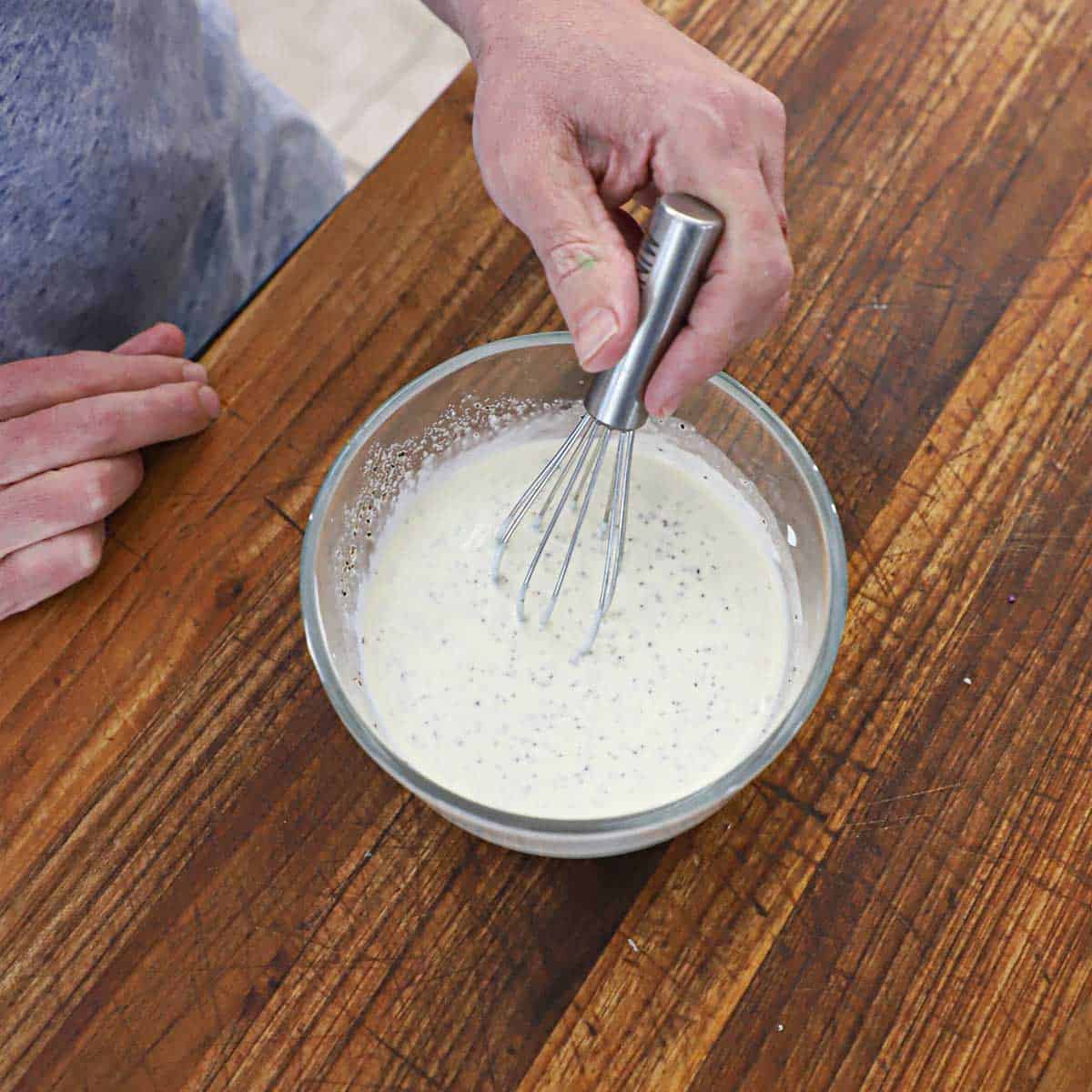 A person whisking a creamy dressing for coleslaw in a small glass bowl sitting on a wooden cutting board.