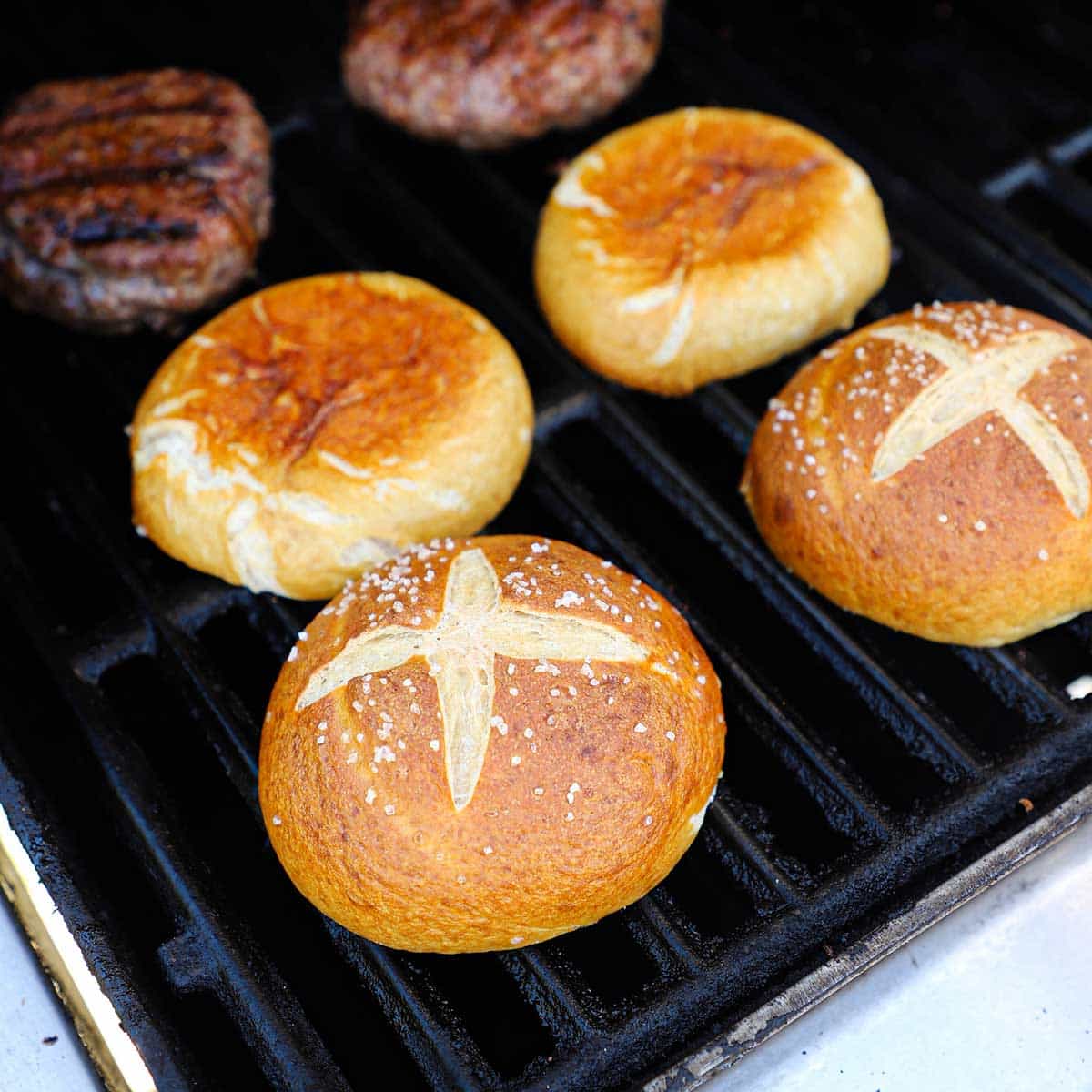 Four pretzel buns that have been cut in half are being toasted, cut-side down, on the grate of a gas grill.
