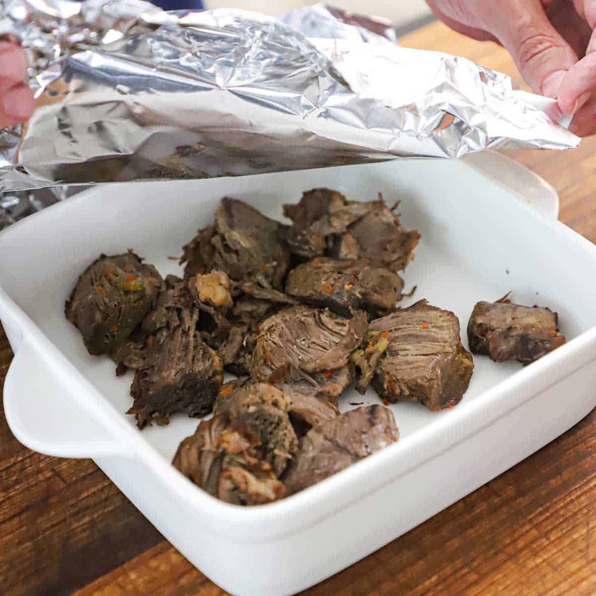 A person placing foil over a square white baking dish that is filled with pieces of fully cooked beef pot roast.