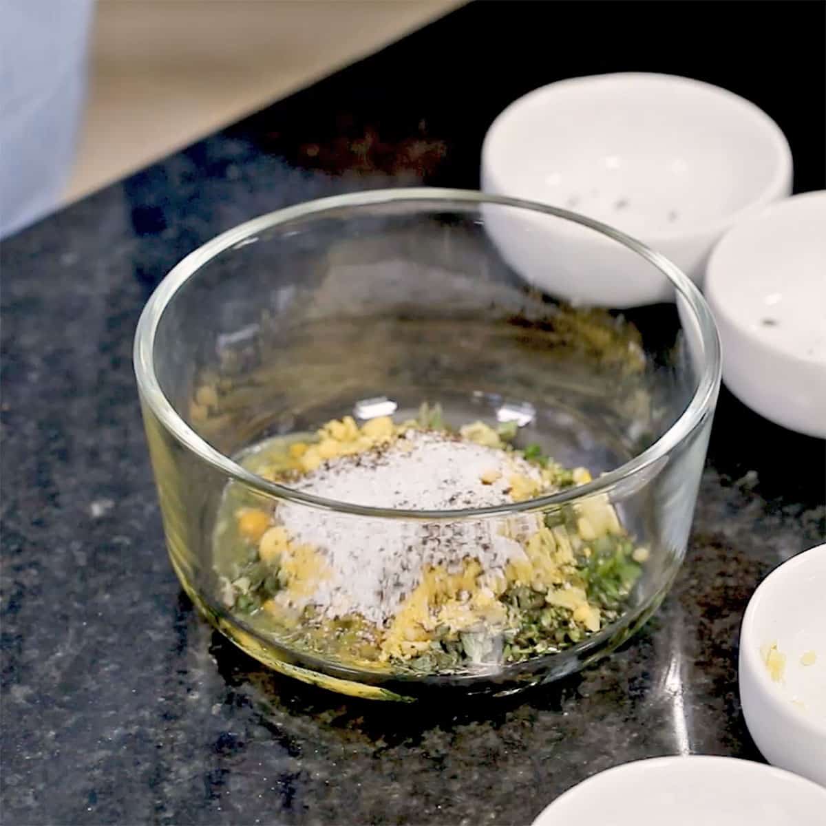 A glass bowl filled with chopped fresh herbs, minced garlic, salt, and pepper.