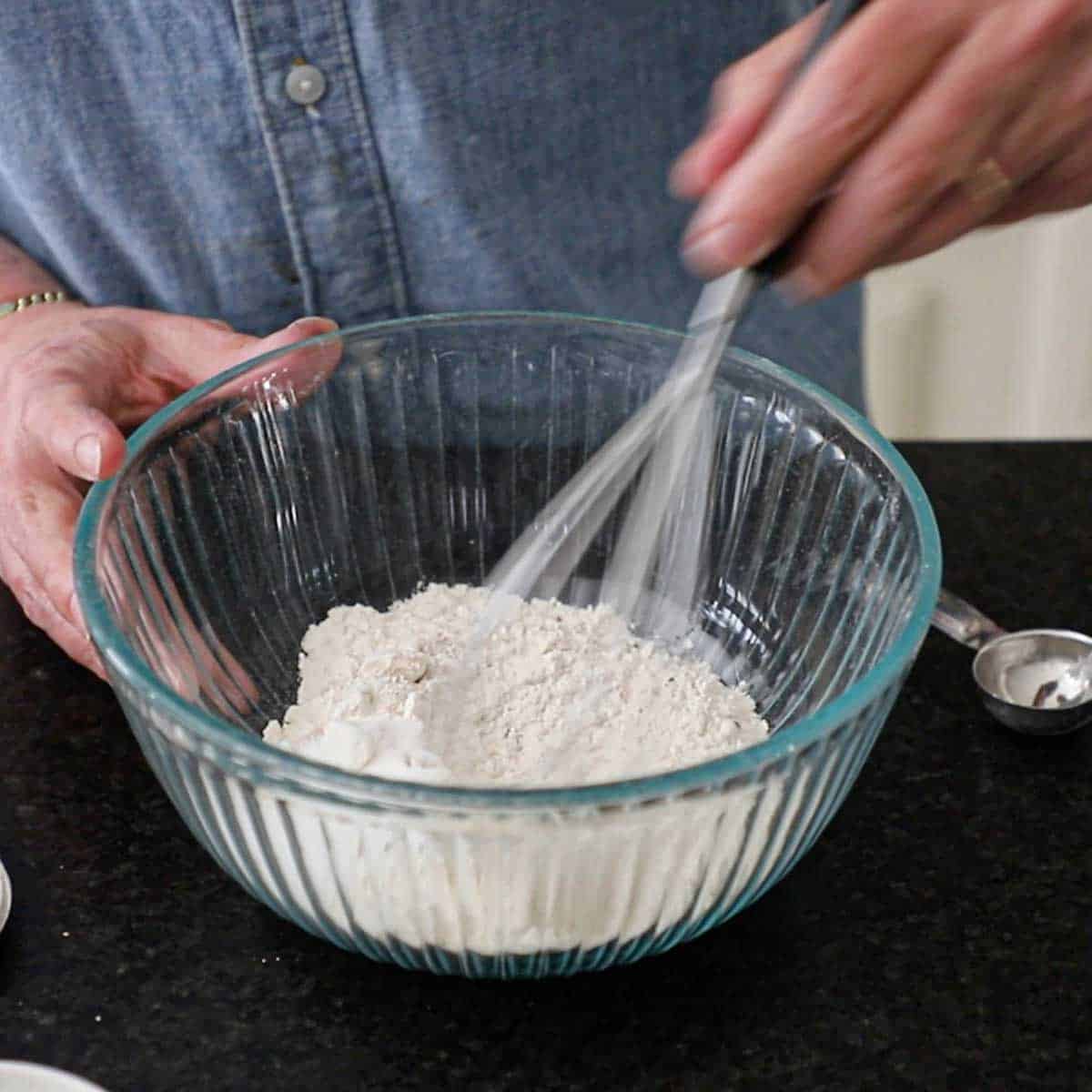 A person using a whisk to mix together high-gluten flour, instant yeast, and salt in a glass bowl.