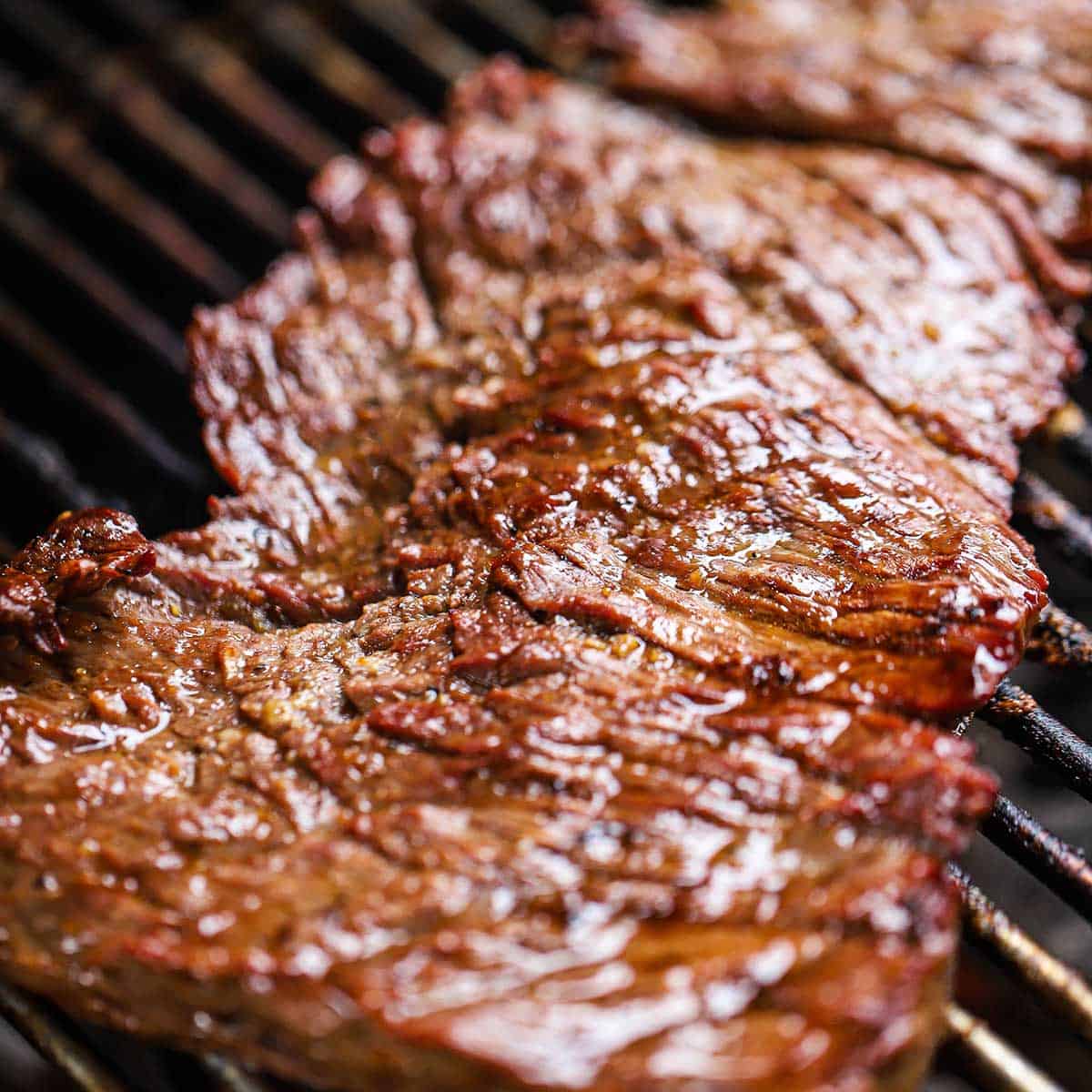 An unsliced strip of seasoned flat iron steak cooking on the grate of a gas grill.