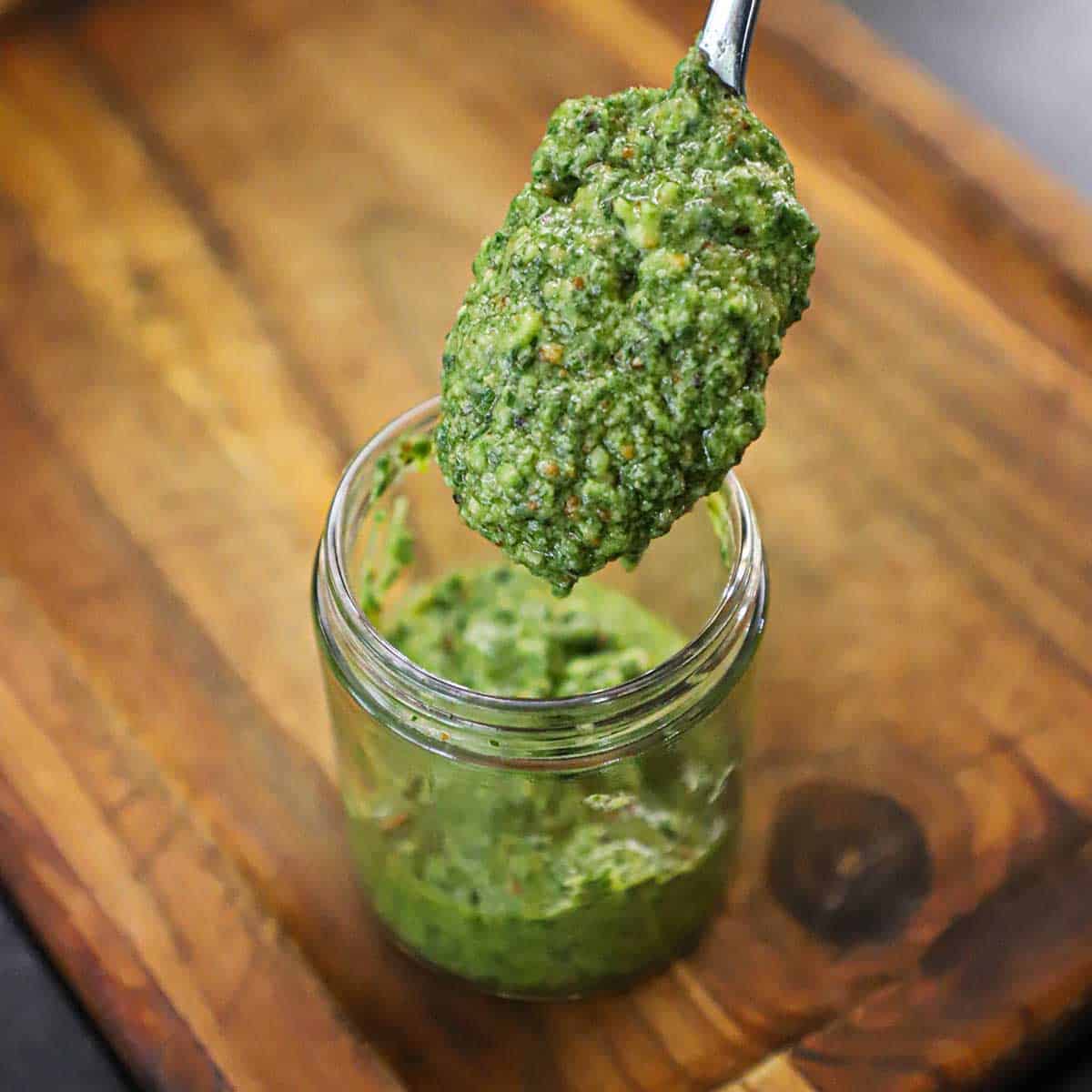 A person holding a heaping spoonful of easy pesto sauce over a small glass jar that is partially filled with the rest of the sauce.