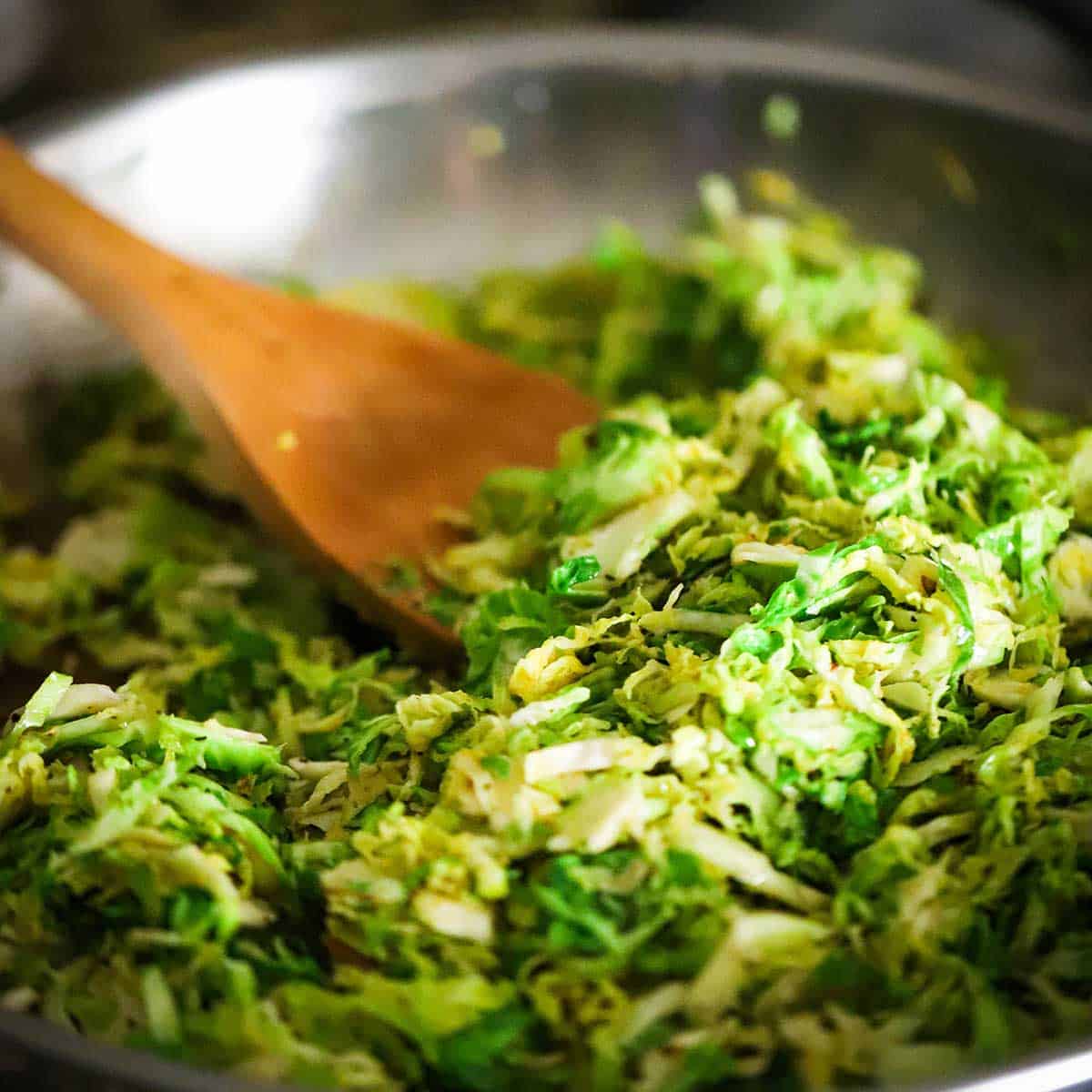 Shave Brussels sprouts being sautéed in white wine in a large silver skillet and being stirred by a wooden spatula.