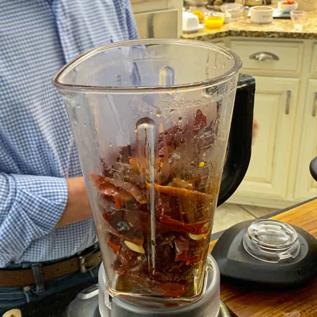 A person standing behind a blender full of softened chiles, garlic, spices, and water on a cutting board on a kitchen island.