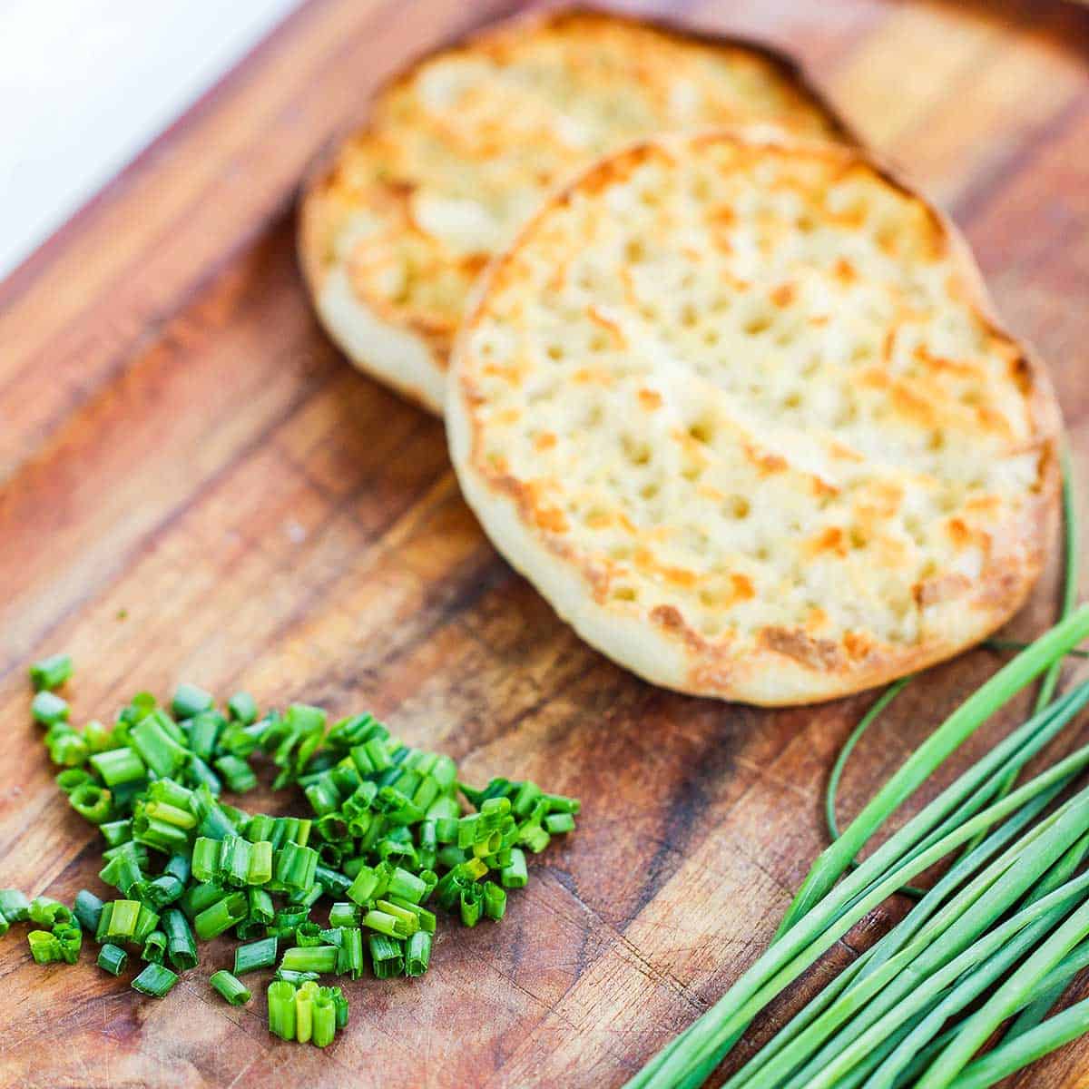 One halved toasted English muffin sitting on wooden cutting board next to a pile of chopped chives and a pile of whole chives.