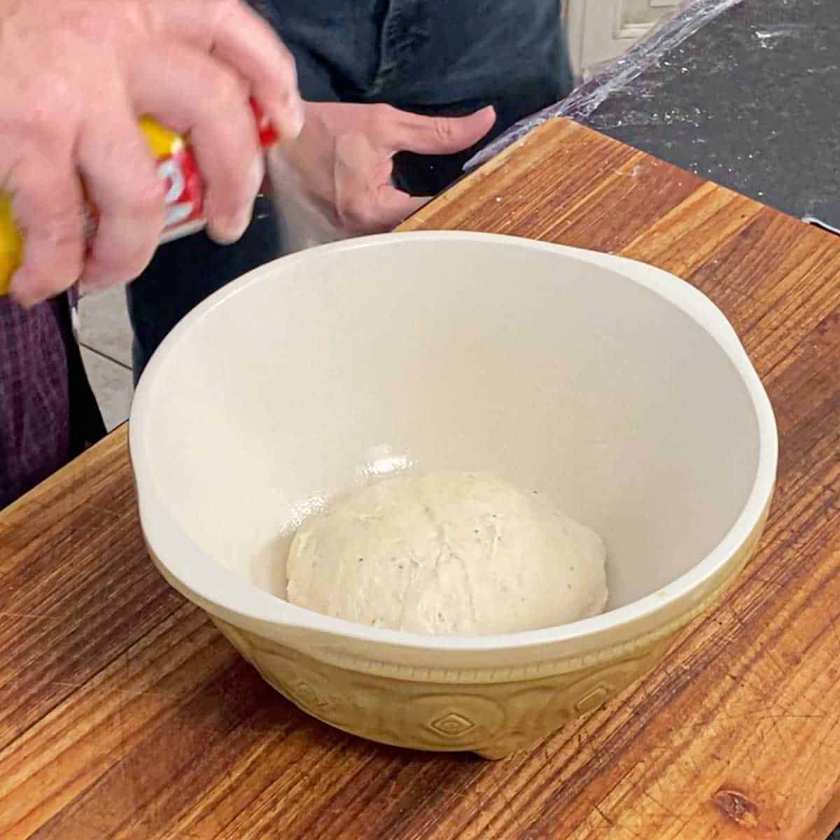 A person spraying cooking spray over an un-risen ball of bagel dough in an oiled ceramic bowl.