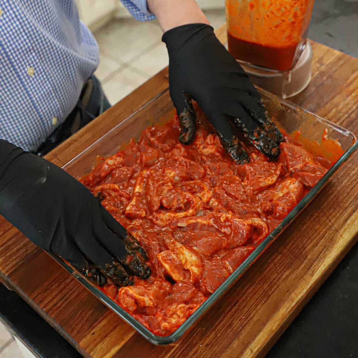 A person wearing latex gloves pressing slices of pork soaked in an al pastor marinade in a large glass baking dish with a blender nearby containing more of the marinade.
