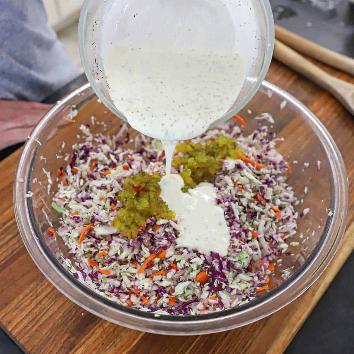 A person pouring a creamy poppyseed dressing into a bowl filled with shredded cabbage, carrots, and sweet dill relish.