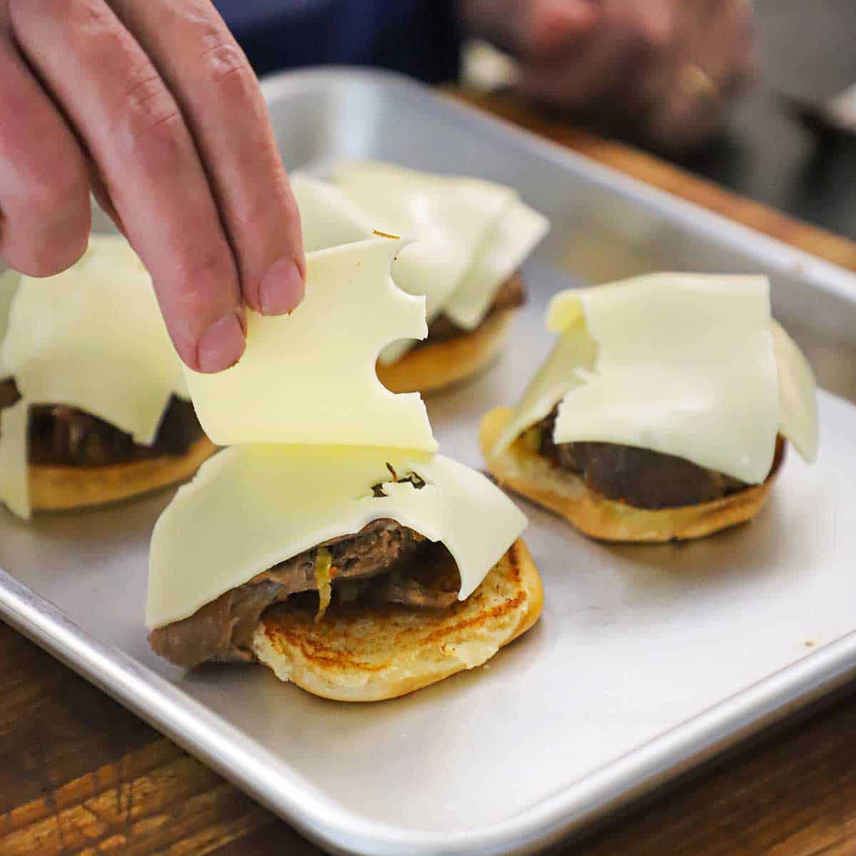 A person placing strips of Swiss cheese over the tops of pot roast that are resting on a the bottom half of a toasted slider bun.