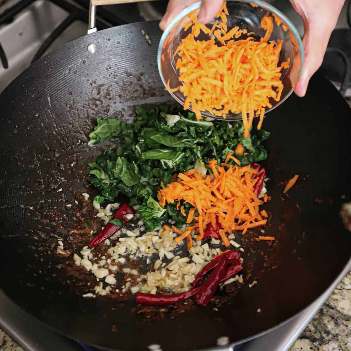 A person dumpling shredded carrots from a small glass bowl into a wok filled with chopped bok choy, garlic, ginger, and chili peppers simmering in oil.