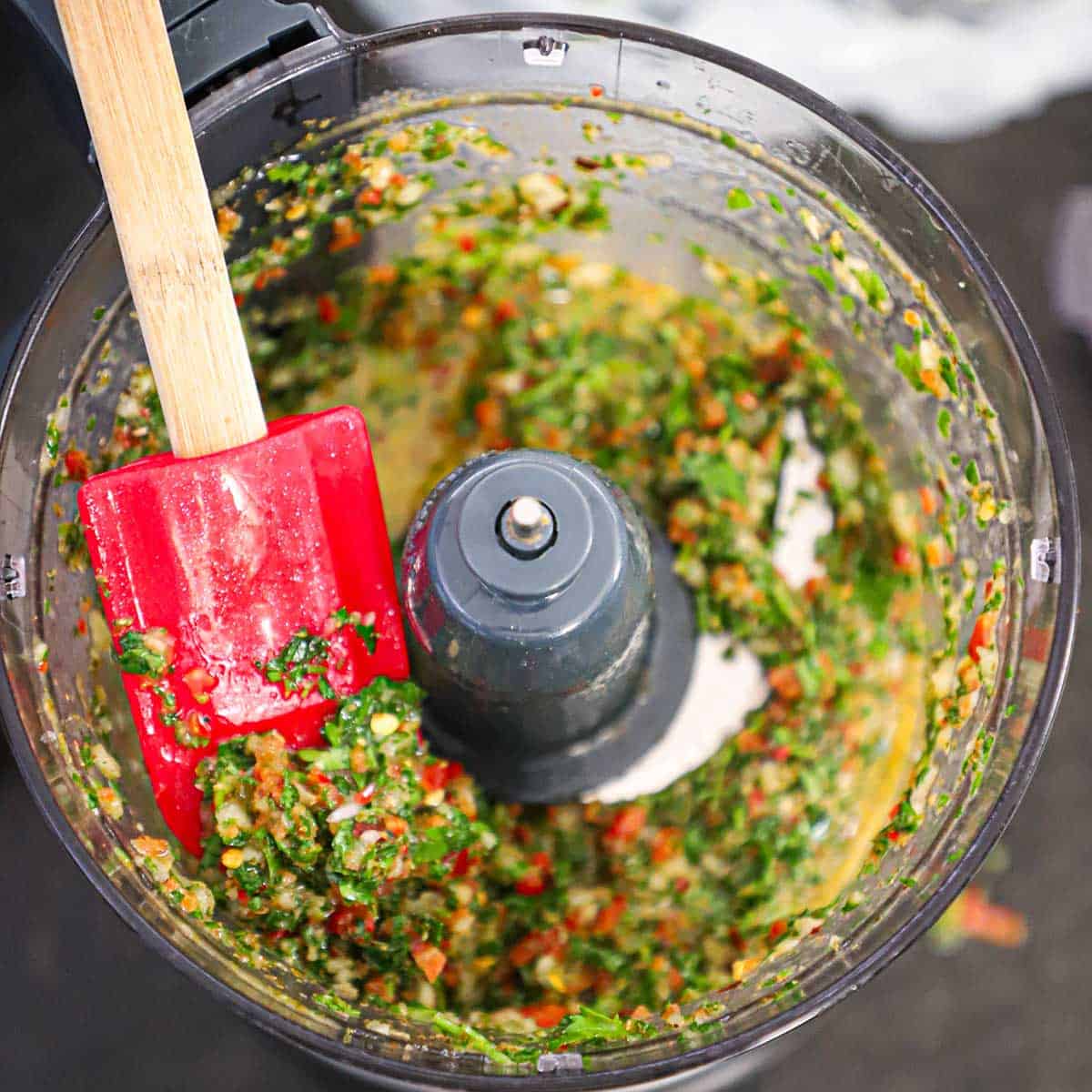A person using a red spatula to hold up freshly chopped chimichurri sauce in the base of a food processor.