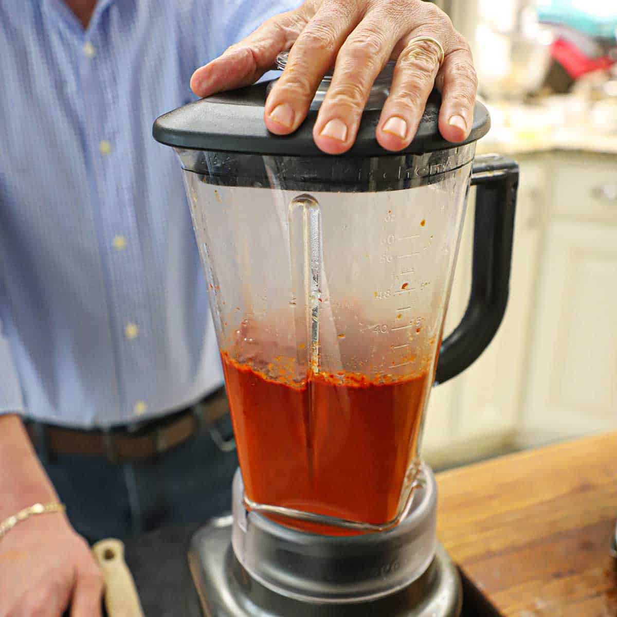 A person holding down the lid of a blender that is halfway filled with a pureed chili mixture that will be a marinade for tacos al pastor.