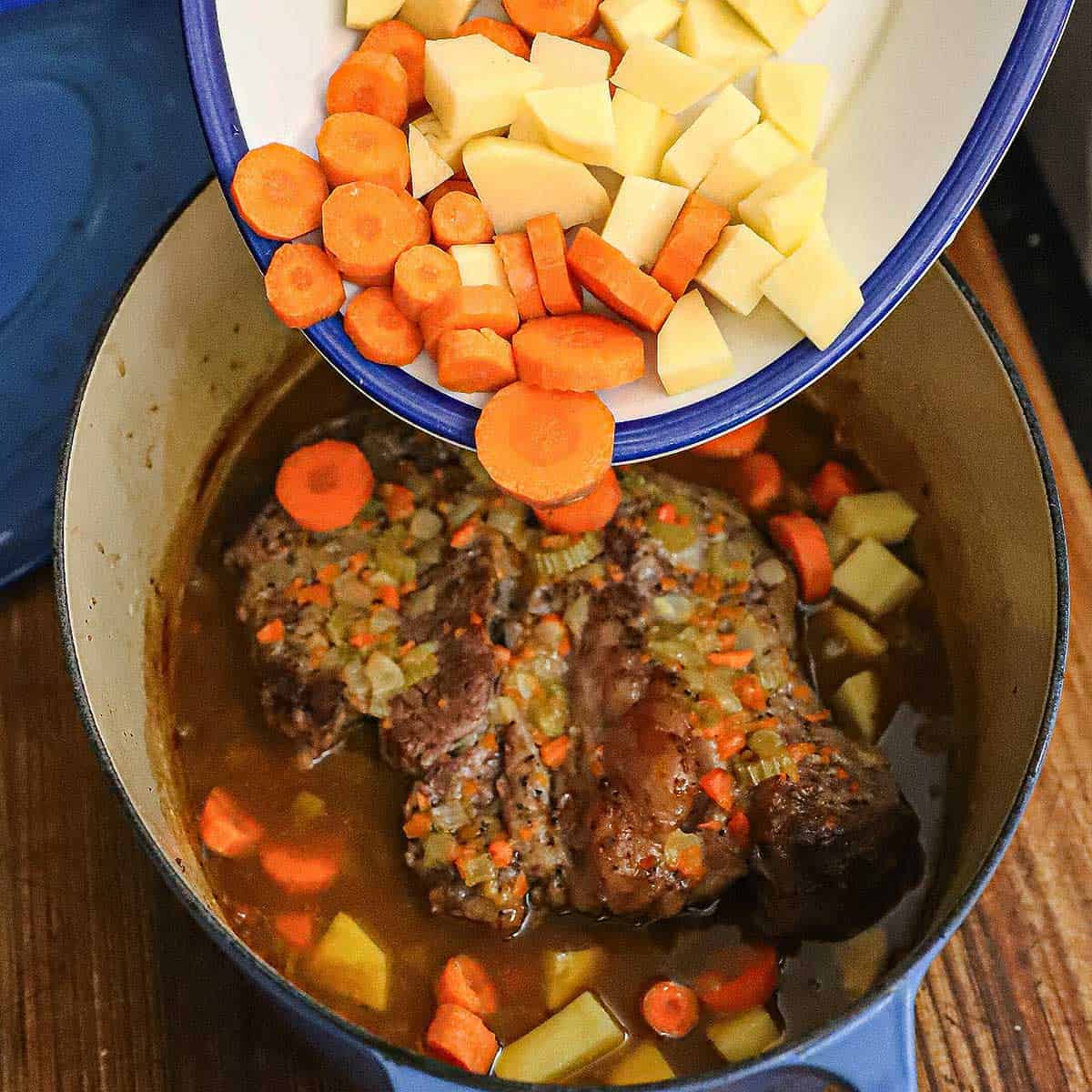 Raw cubes of white potatoes and slices of raw carrots being transferred from a platter into a Dutch oven filled with a partially cooked chuck roast, beef broth, and sautéed mirepoix.