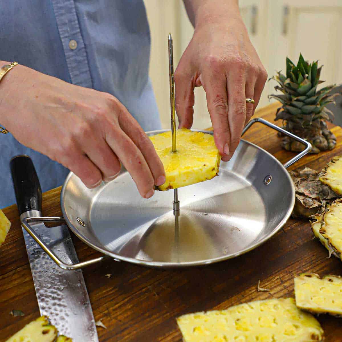 A person pressing a large slice of fresh pineapple onto an upright skewer that is attached to a metal pan with handles on a wooden cutting board.