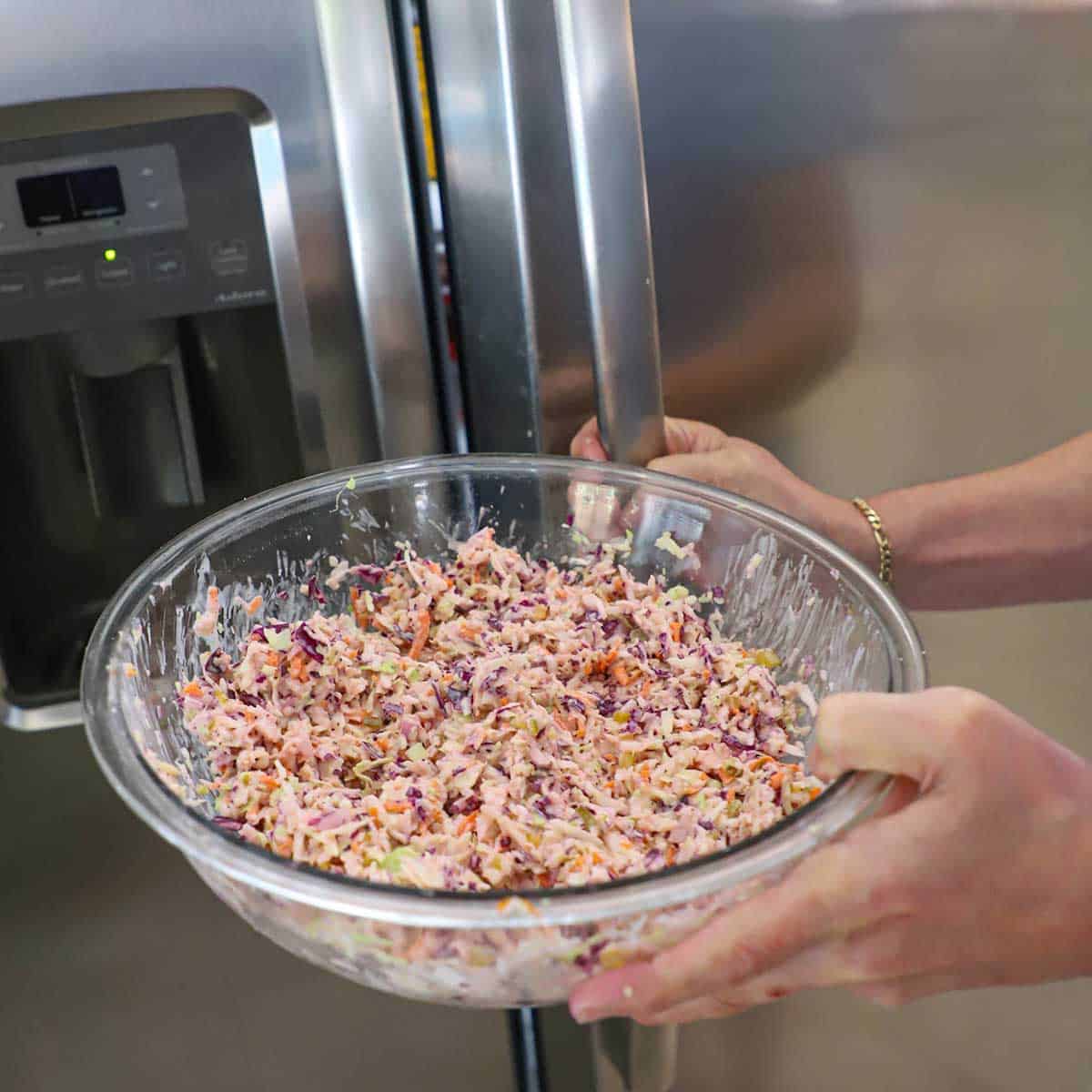 A person opening a refrigerator door with one hand and holding a glass bowl filled with diner-style coleslaw in the other hand.