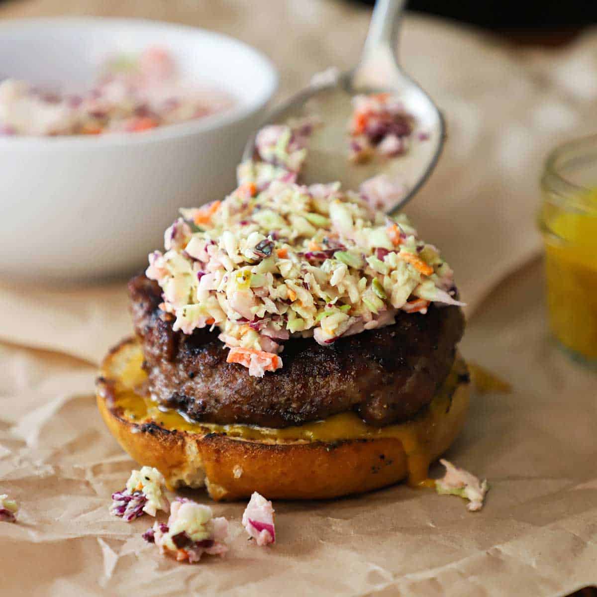 A person using a spoon to add diner-style coleslaw on the top of a grilled pork burger that is resting on the bottom half of a pretzel bun with mustard sauce on it.