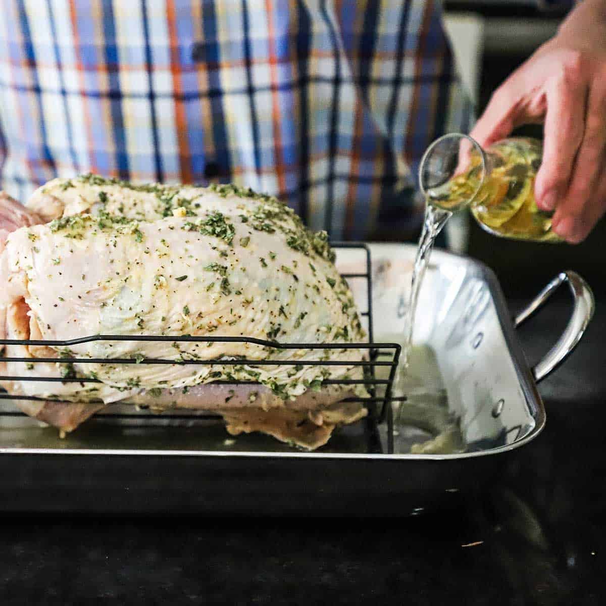 A person pouring white wine from a small carafe into a roasting pan that is fitted with a roasting rack with an uncooked turkey breast with herbs on it.
