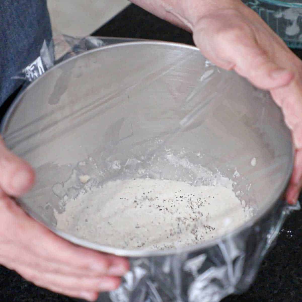 A person holding the bowl of a stand mixer filled with a bagel dough sponge with clear plastic wrap over the top.