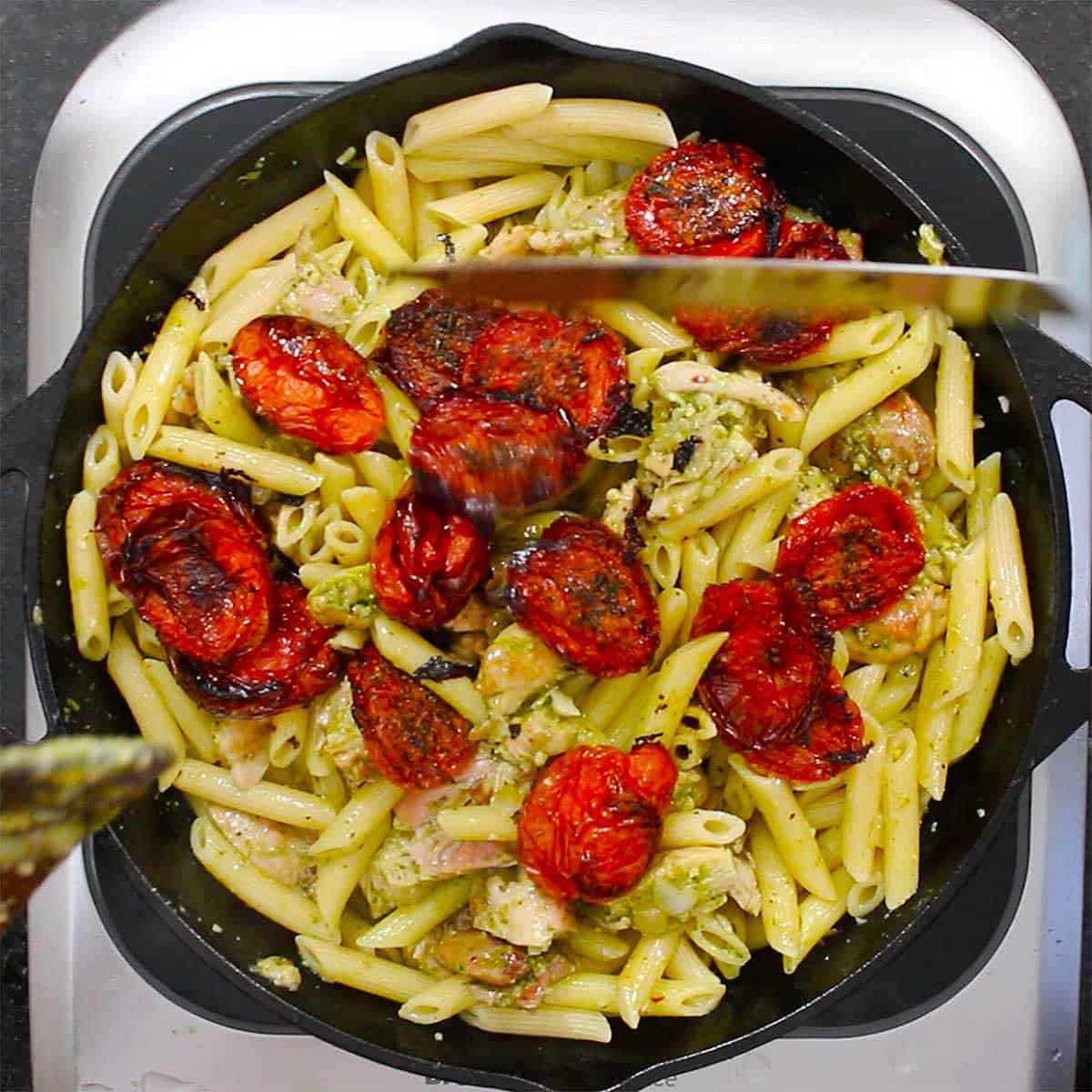 A large black cast-iron skillet filled with cooked pasta, pesto chicken and roasted Roma tomatoes.