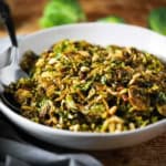 A white serving bowl filled with shaved Brussels sprouts with balsamic, pine nuts, and pancetta with a large spoon inserted into the side of the salt.