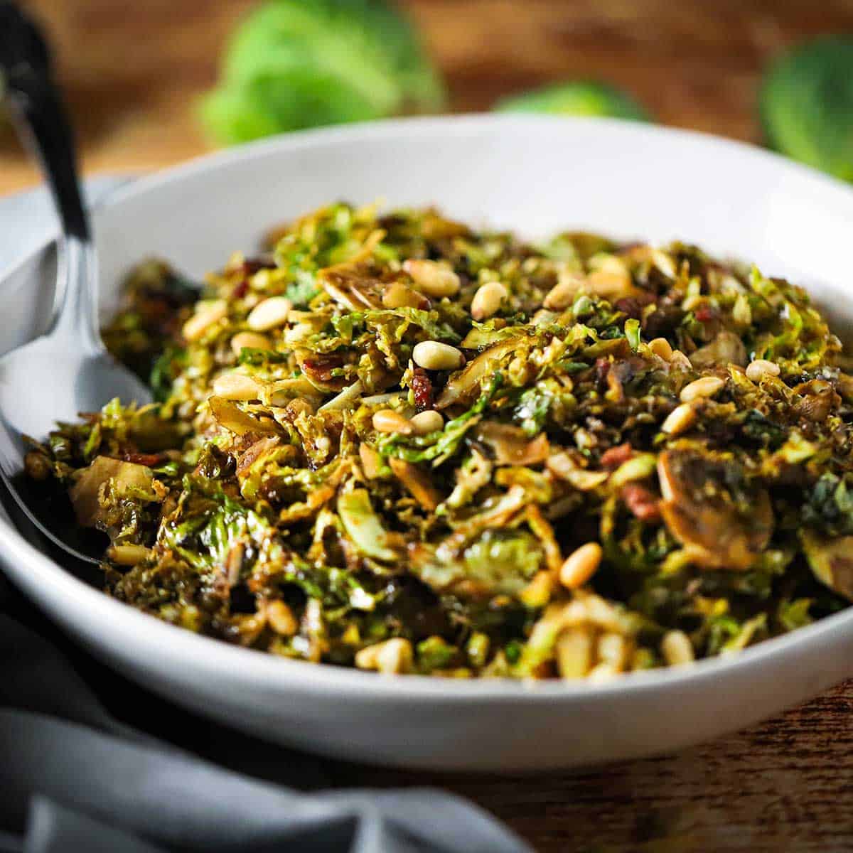 A white serving bowl filled with shaved Brussels sprouts with balsamic, pine nuts, and pancetta with a large spoon inserted into the side of the salt.