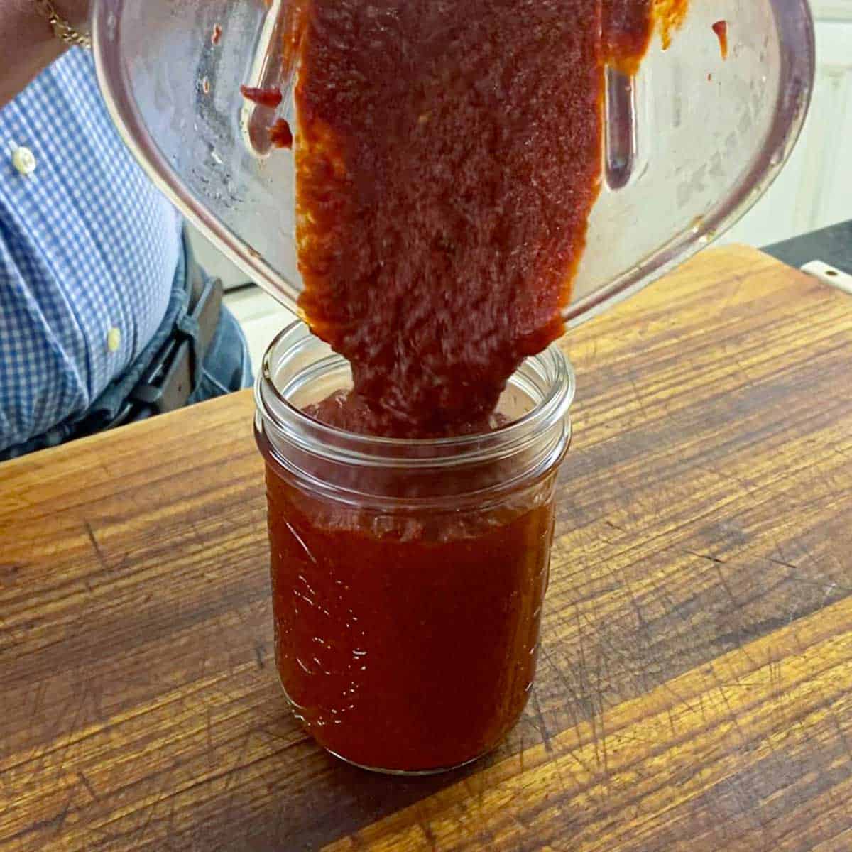 A person pouring a bright red marinade from a blender into a glass jar on a wooden cutting board.