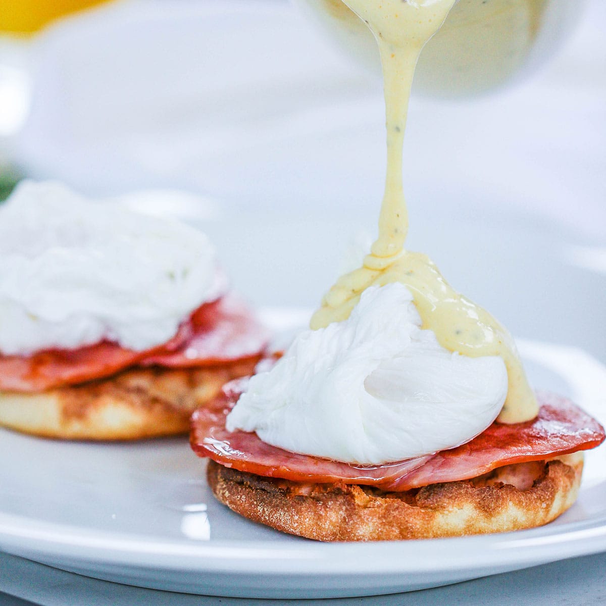 Hollandaise sauce being poured from a glass gravy boat over a poached egg resting on crispy Canadian bacon and a toasted English muffin on a white plate.