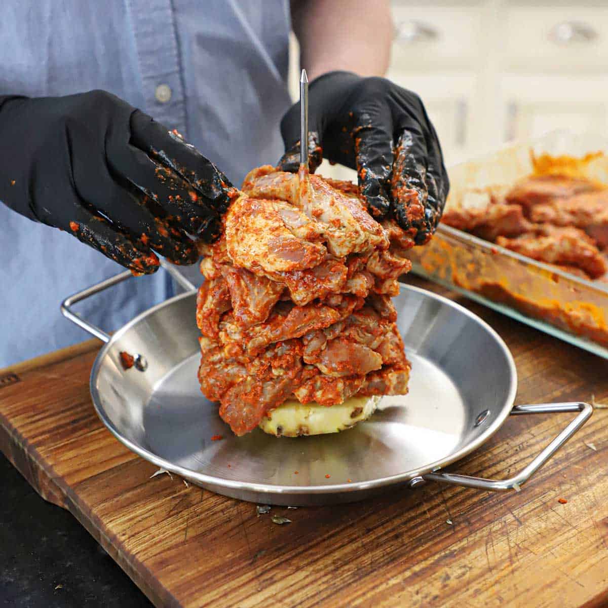 A person pressing onto the top of a pile of strips of raw marinated pork shoulder on an upright skewer attached to a metal pan with handles.