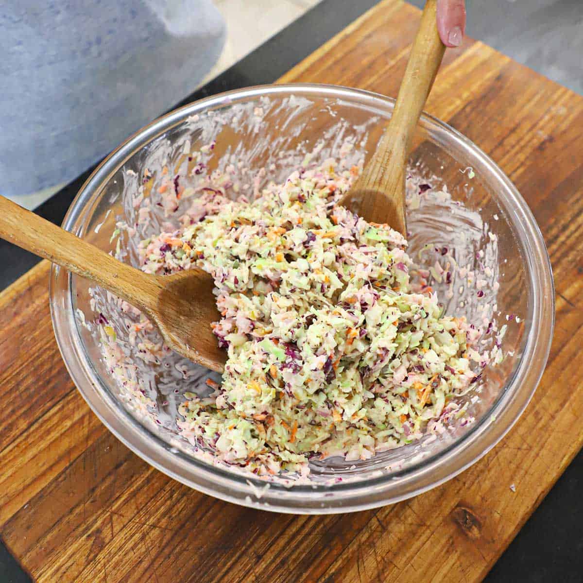 A person using two large wooden spoons to toss diner-style coleslaw in a large glass bowl.