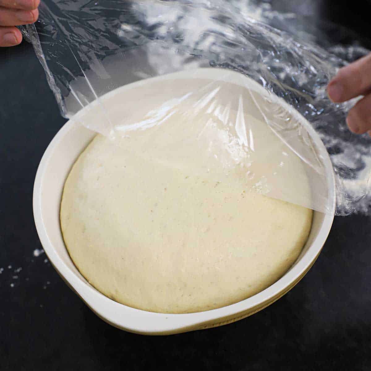 A person removing plastic wrap the top of a ceramic bowl that is filled with risen pretzel dough.