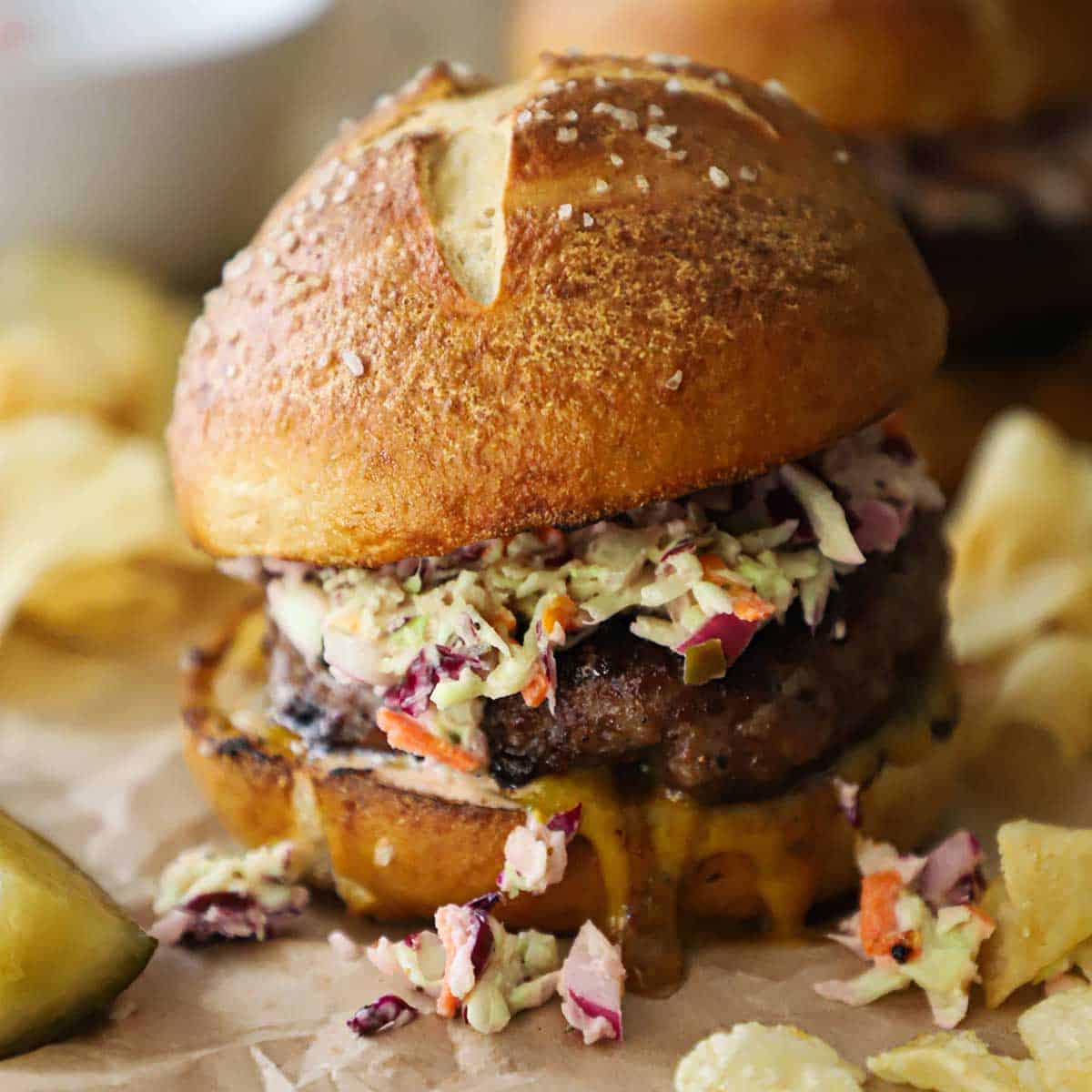 A pork burger topped with creamy coleslaw on a homemade pretzel bun with Carolina mustard sauce oozing from the underneath the patty with kettle chips scattered around the burger.