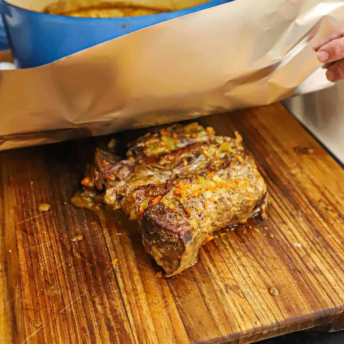 A person placing a large piece of aluminum foil over the top of a fully cooked classic pot roast that is resting on a wooden cutting board.
