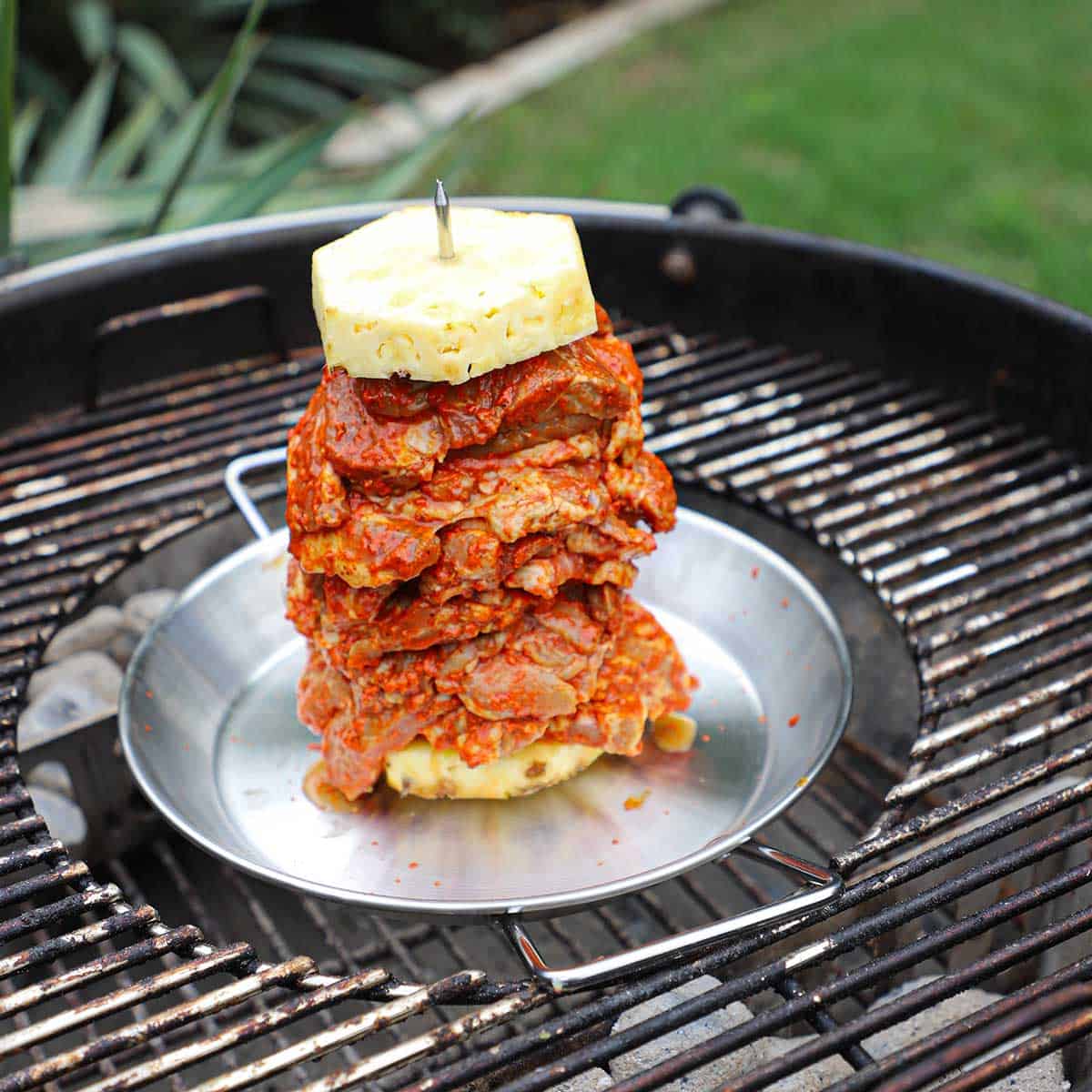 A stack of pork with pineapple slices on an al pastor pan resting in the middle of a charcoal kettle grill.