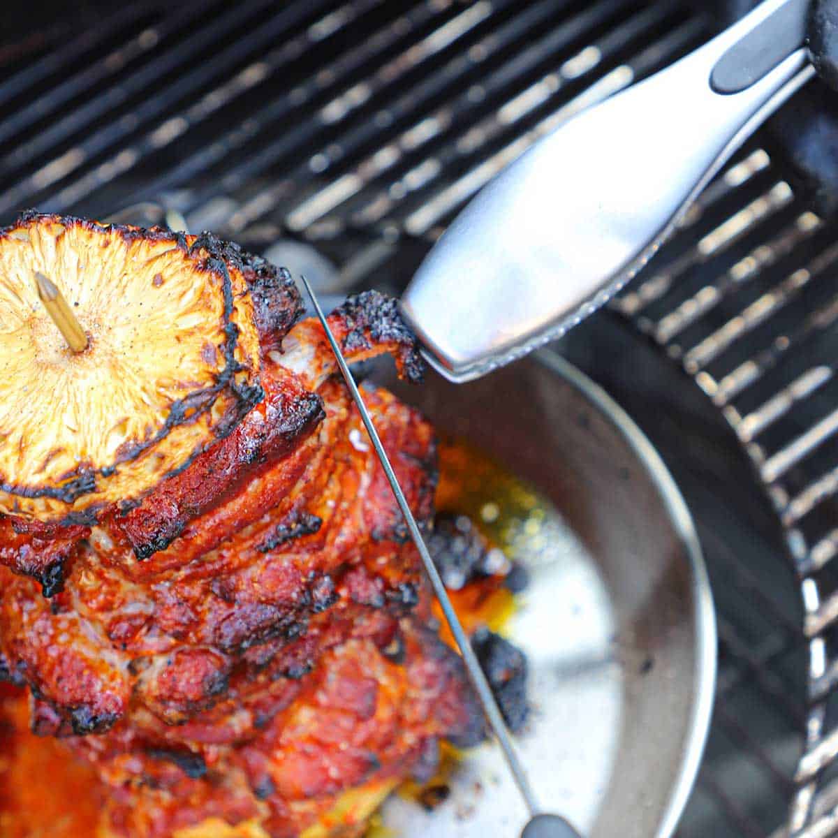A person using a chef's knife and a pair of tongs to cut away burnt ends of a partially cooked pork al pastor on a charcoal grill.