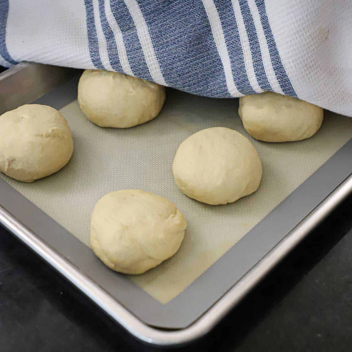A kitchen towel being lifted away from a baking sheet that is lined with a silicone matt and filled with round balls of pretzel dough.