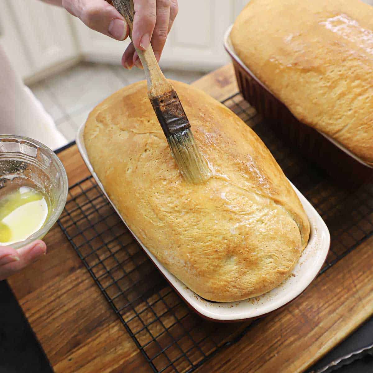 A person using a pastry brush to brush melted butter over the top of a freshly baked loaf of country white bread.
