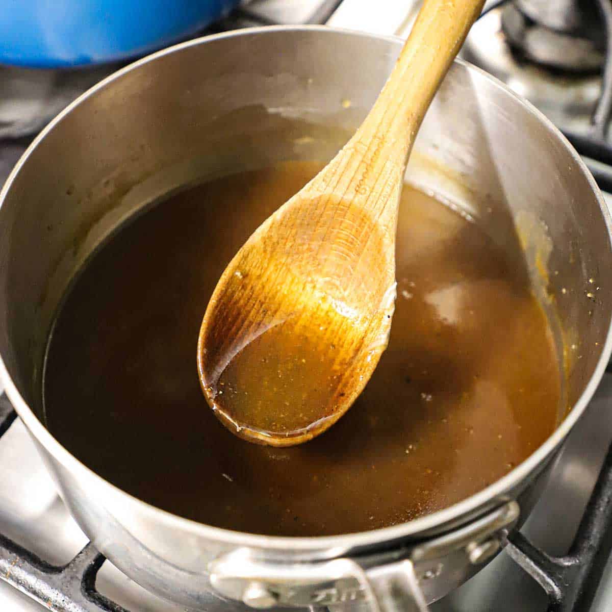 A person using a wooden spoon to lift a small amount of brown gravy from a medium-sized saucepan filled with the gravy.