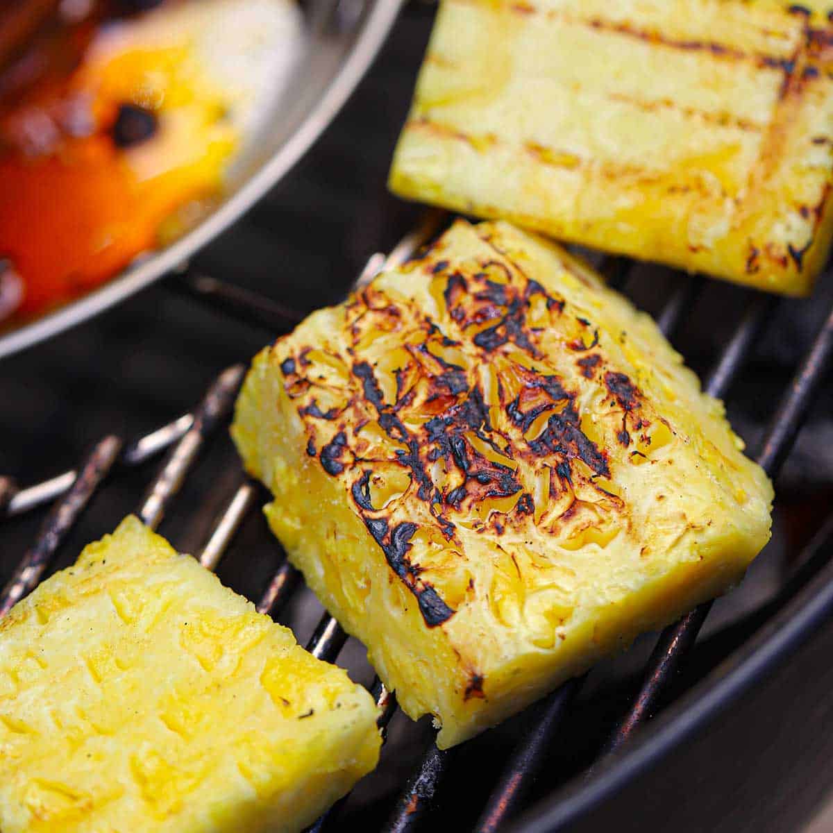 Several large grilled chunks of fresh pineapple resting on the edge of a grate on a charcoal grill.